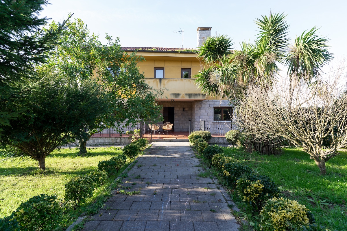 The exterior of the chalet is surrounded by well-maintained gardens featuring shrubs and palm trees. A paved pathway leads to the entrance, which is flanked by a covered porch. A spacious balcony extends from the upper level, offering views of the lush greenery.