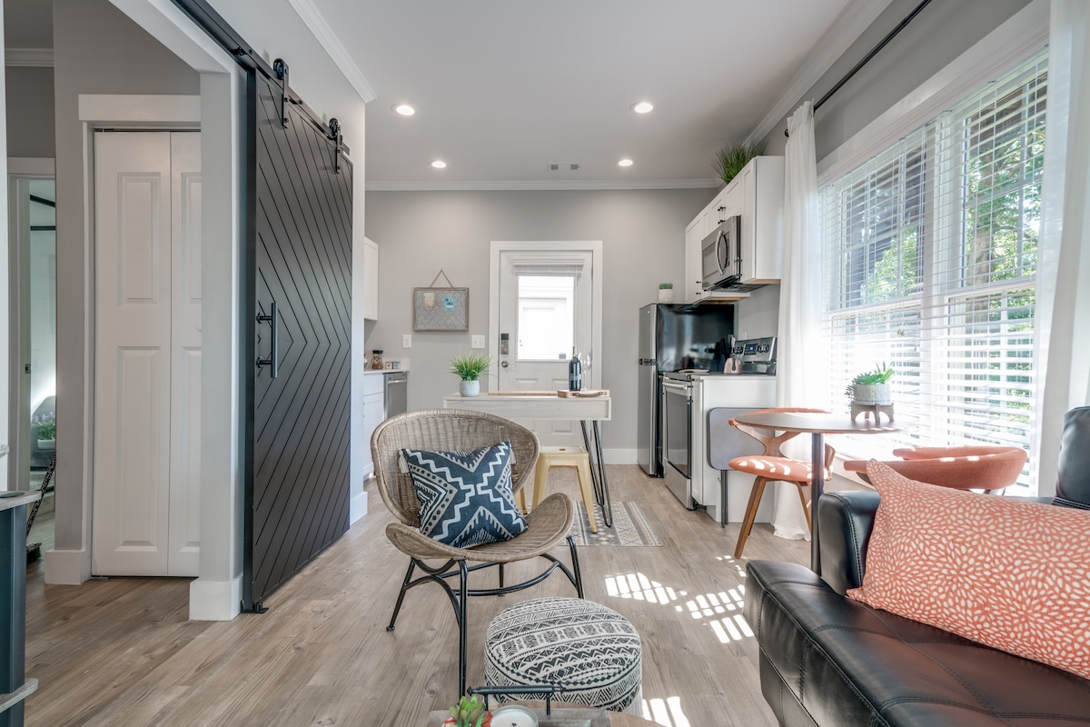 An inviting living space features a combination of modern furniture and natural light. A comfortable seating area includes a black leather couch, a patterned accent chair, and a small coffee table. A barn door adds a touch of rustic charm, while the kitchenette is visible in the background.