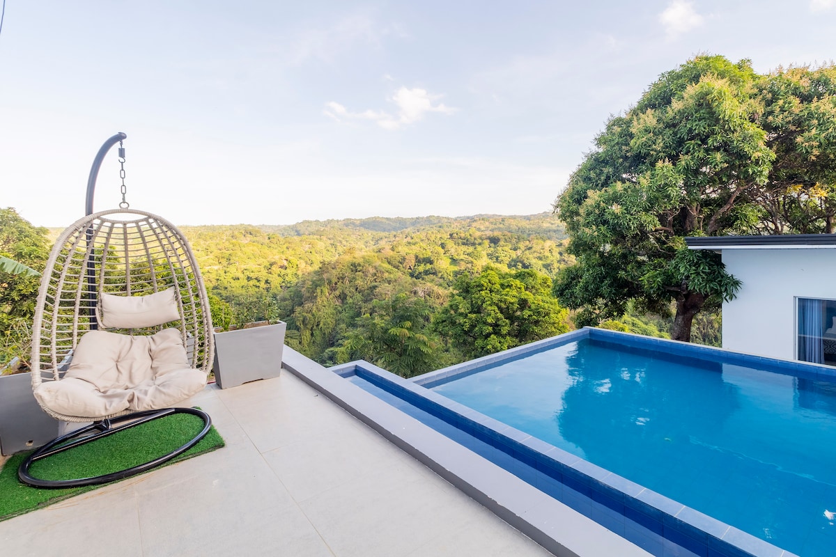 An outdoor space features a hanging chair beside an infinity pool with serene views of lush greenery. The tranquil water reflects the sky, while the surrounding landscape is filled with trees, creating a peaceful atmosphere for relaxation.