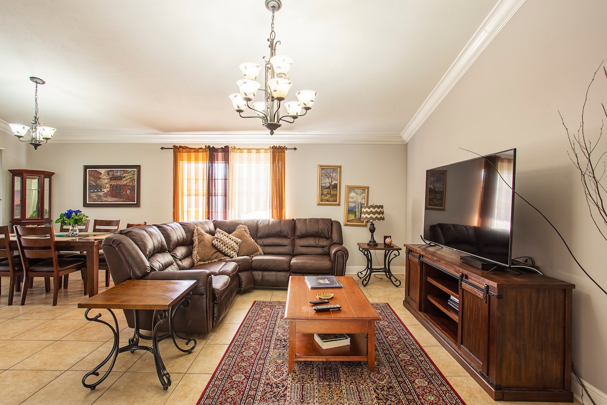 A spacious living area features a large sectional sofa complementary to a wooden coffee table. A flat-screen TV is positioned on an entertainment unit, while a dining table is placed in the background. Natural light filters through the window, accentuated by light curtains.
