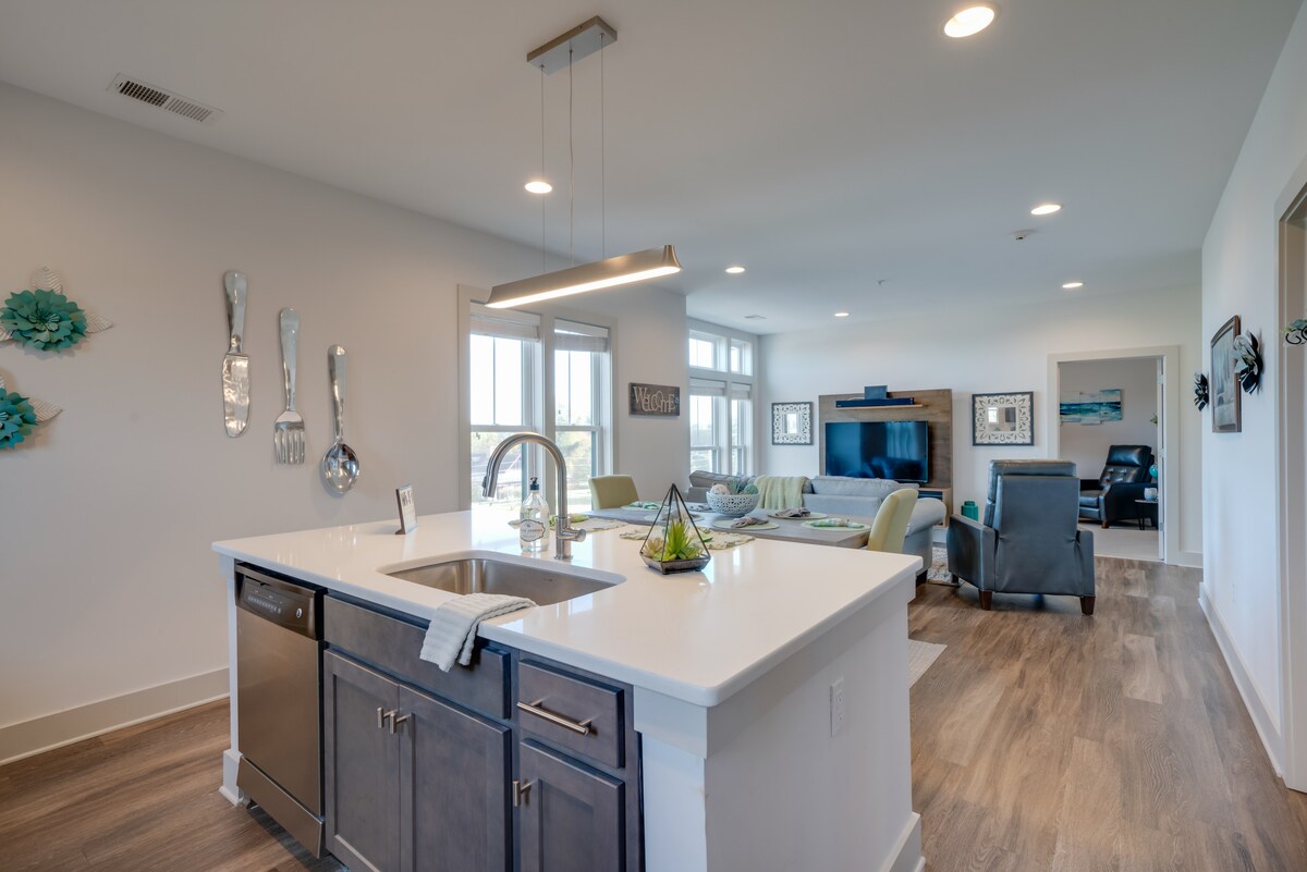 A modern kitchen is displayed with a large island featuring a stainless steel sink and a pendant light above. Open to the living space, large windows allow natural light to flood in, highlighting a comfortable seating area with a flat-screen TV.