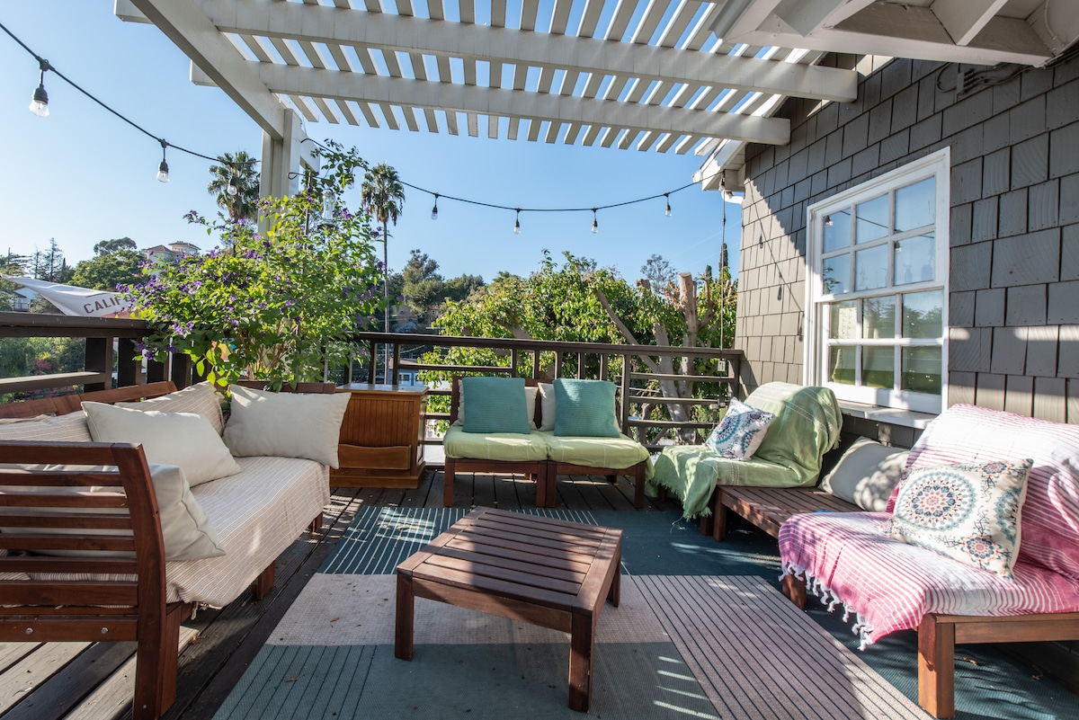 A spacious deck area is furnished with comfortable seating, including a mix of sofa and lounge chairs. A wooden table is positioned in the center, surrounded by greenery and soft lighting from overhead string lights.