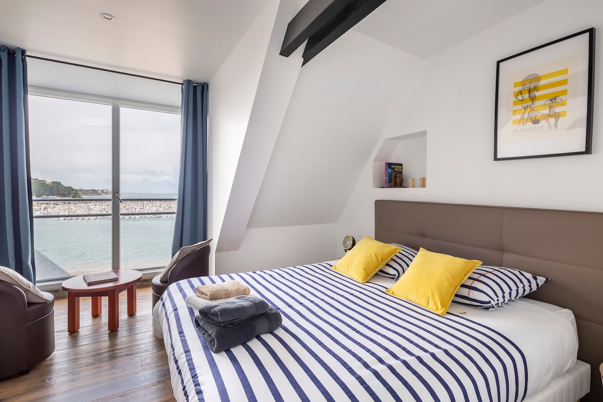 A spacious sleeping area features a large bed with striped bedding complemented by two bright yellow pillows. An armchair and a small table are positioned near a wide window, offering views of the port and the sea, while natural light brightens the room.