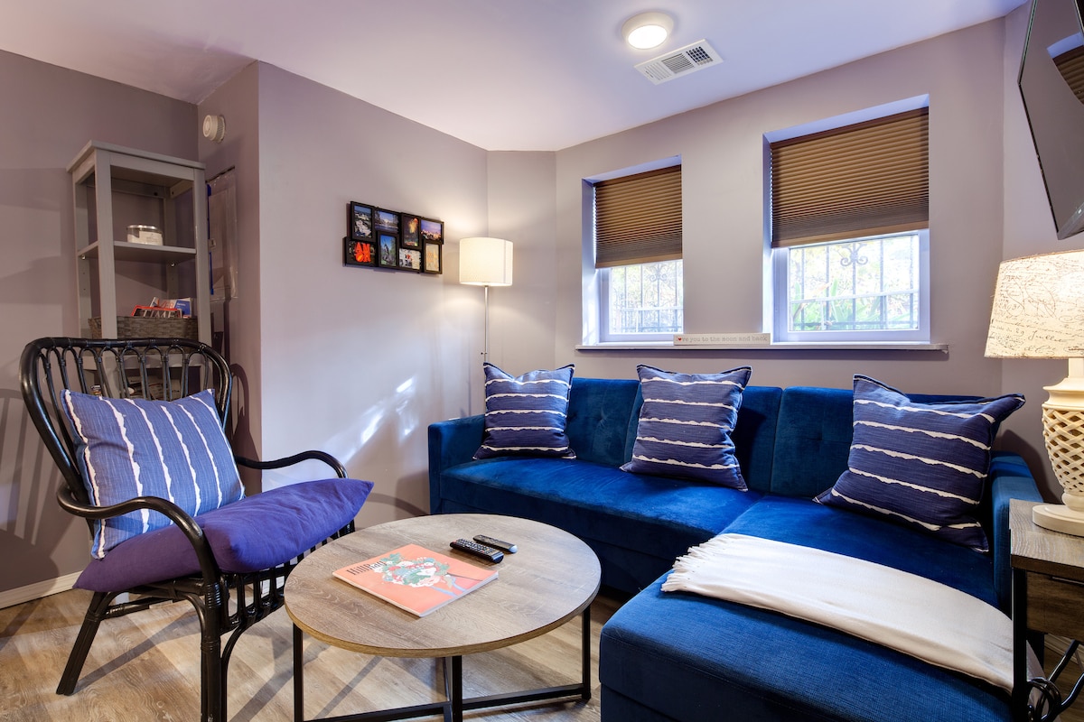 The living area features a comfortable blue sectional sofa with decorative pillows and a cozy wicker chair. A round coffee table sits in the center, surrounded by natural light from two windows with blinds. A wall-mounted TV is mounted to one side.