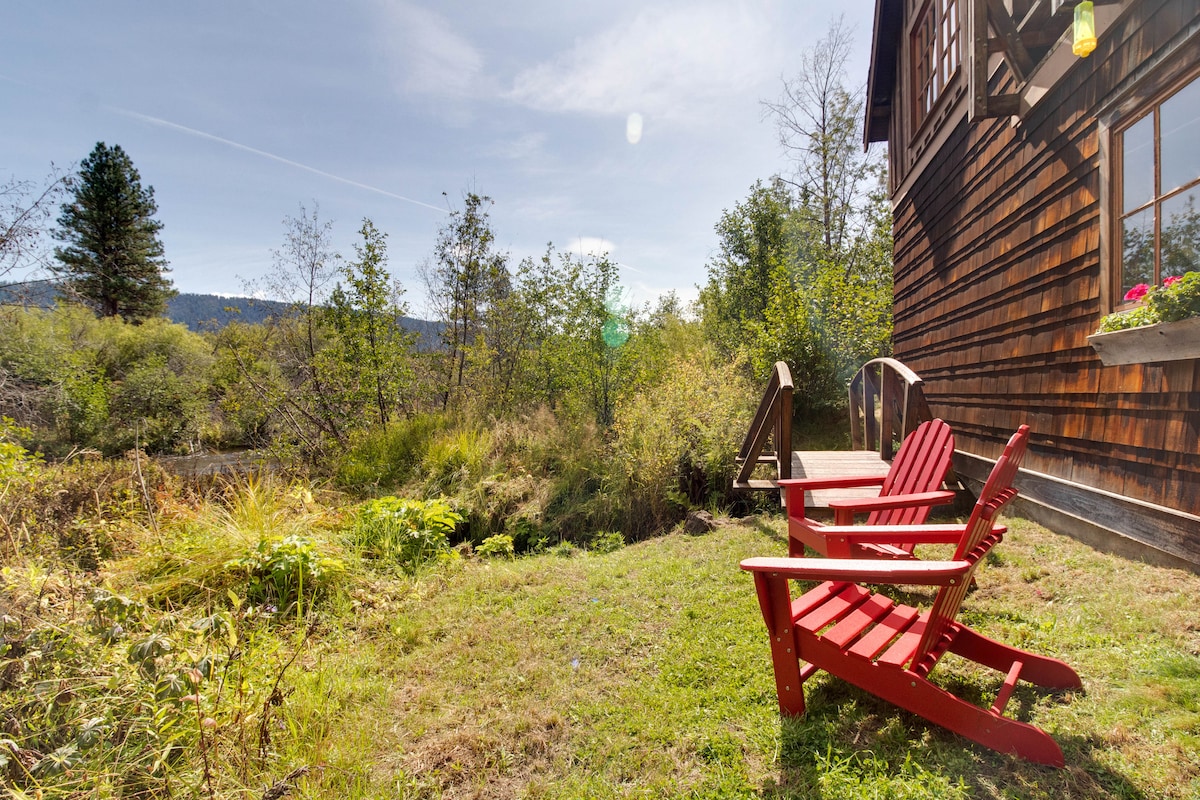 A small outdoor seating area features a pair of red Adirondack chairs positioned on a grassy patch. Surrounding the area, native vegetation and trees create a natural backdrop, while the distant landscape is visible under a clear blue sky.