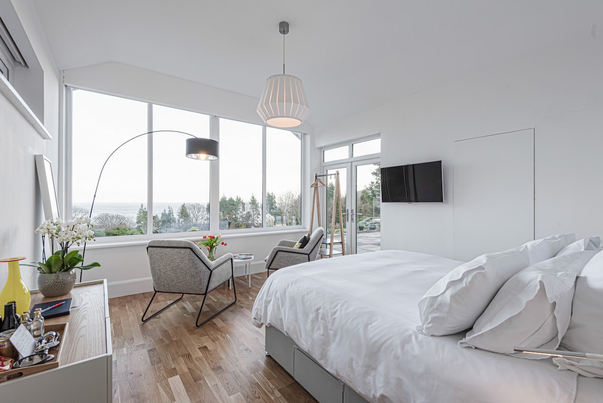 A spacious bedroom features a super king size bed, flanked by two comfy chairs positioned by large windows, showcasing stunning views. A flat-screen smart TV is mounted on the wall. Natural light fills the room, enhancing the modern decor and wooden flooring.