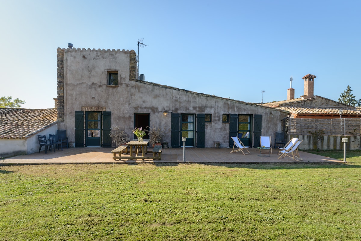 The exterior of the house is characterized by rustic architecture, with a textured stone facade and multiple windows. A spacious terrace is visible, featuring outdoor seating and a wooden table. Lush green grass surrounds the house, enhancing the inviting outdoor space.