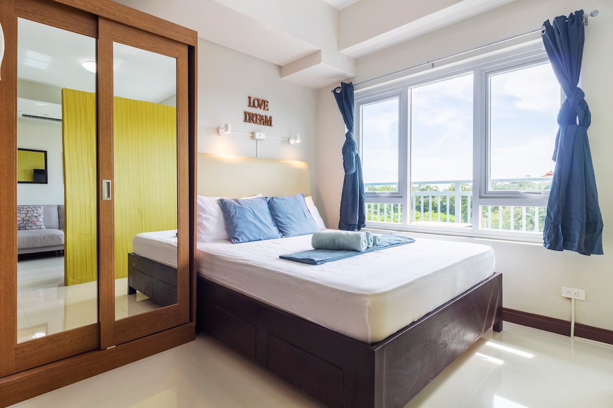 A bright sleeping area is equipped with a spacious bed dressed in fresh linens and two accent pillows. A large mirror reflects natural light from the window, which is framed by dark blue curtains. An inspirational quote is displayed on the wall above the bed.