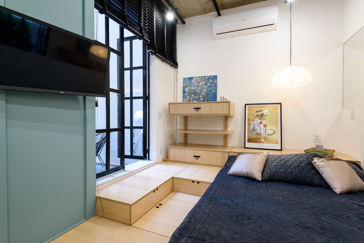 A spacious sleeping area features a large bed with a dark quilt and decorative pillows. A minimalist wooden platform with storage is positioned beside the bed. A wall-mounted TV is visible along with light streaming through multiple black-framed windows. A soft pendant light adds illumination.
