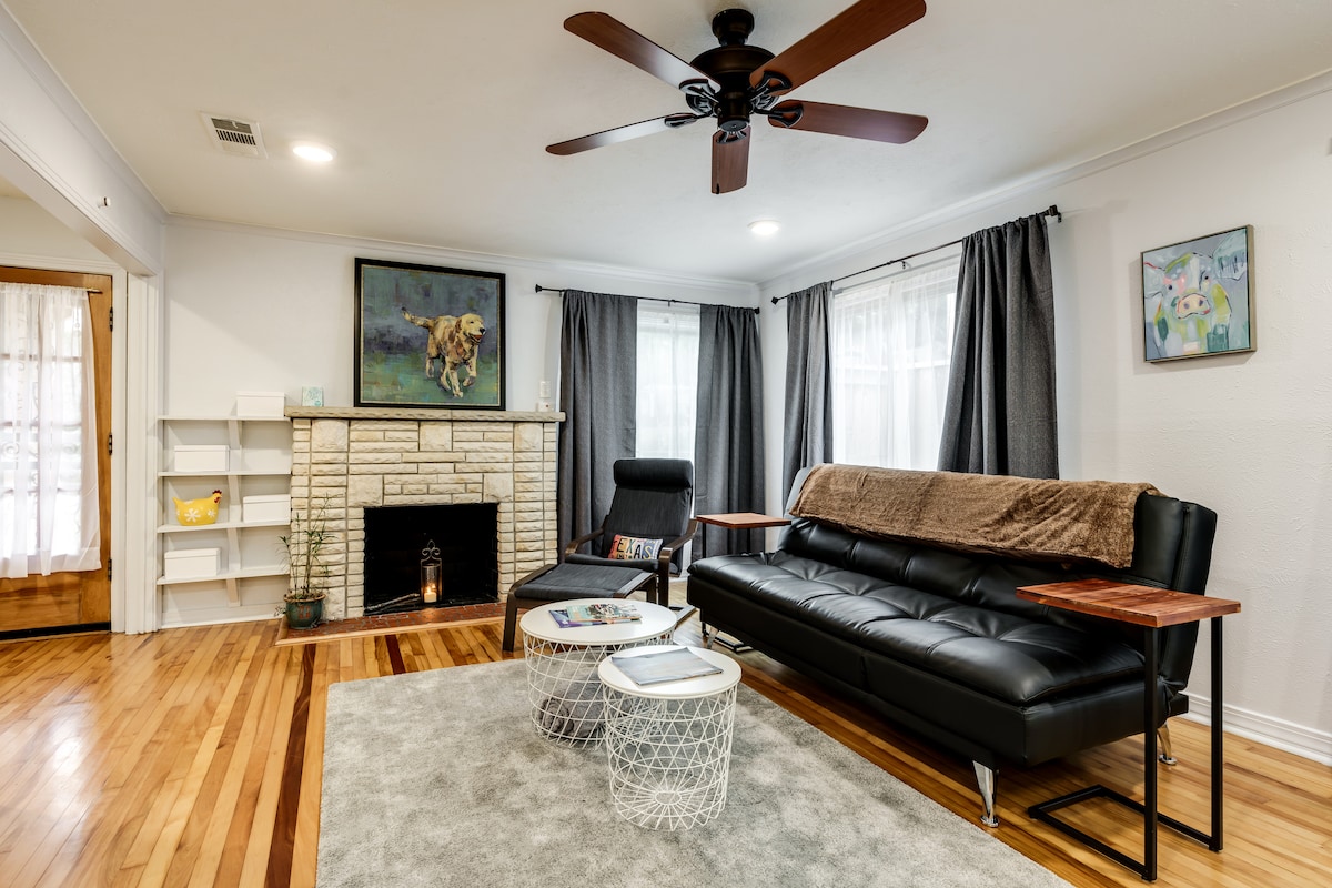 A living area features a sleek black sofa facing a traditional fireplace. Nearby, a stylish black chair is accompanied by two modern side tables. Large windows draped in dark curtains allow natural light to fill the space, adding warmth to the hardwood flooring.