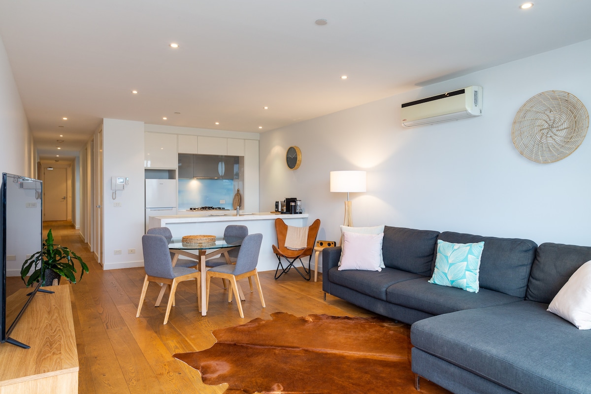 An open-plan living area features a large grey sectional sofa accompanied by a wooden coffee table. A round dining table with four chairs is visible, along with a fully equipped kitchen area. Natural light fills the space, highlighting the clean layout and modern decor.