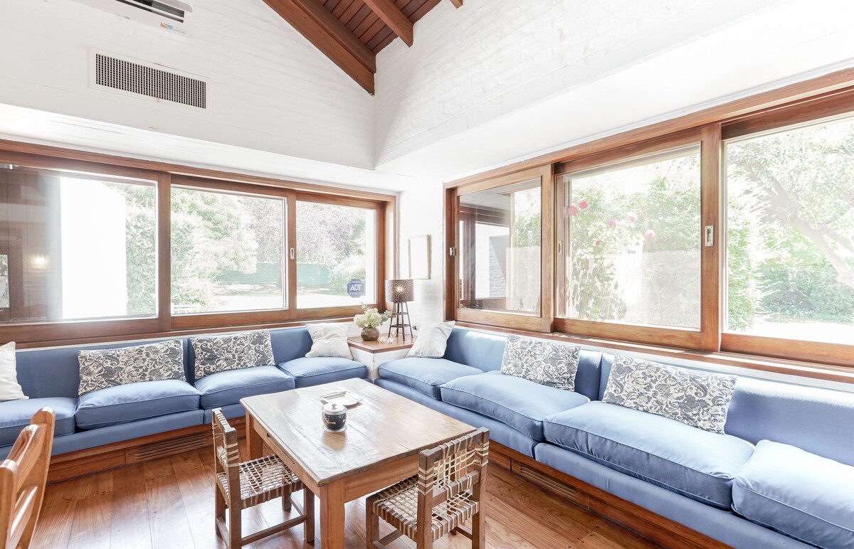 A light-filled living area is featured, showcasing large windows that provide views of the garden. Soft blue sofas surround a wooden table, complemented by a neutral decor that enhances the serene environment. Natural light illuminates the space, creating a welcoming atmosphere.