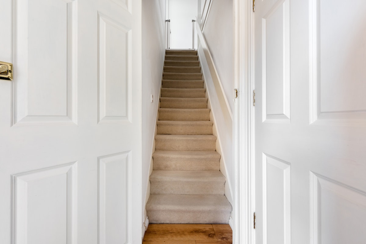 A softly carpeted staircase leads upward, flanked by white panelled walls. Natural light filters in from the upper landing, illuminating the warm wooden floor at the base of the stairs. Double doors are positioned on either side, creating a welcoming entryway.