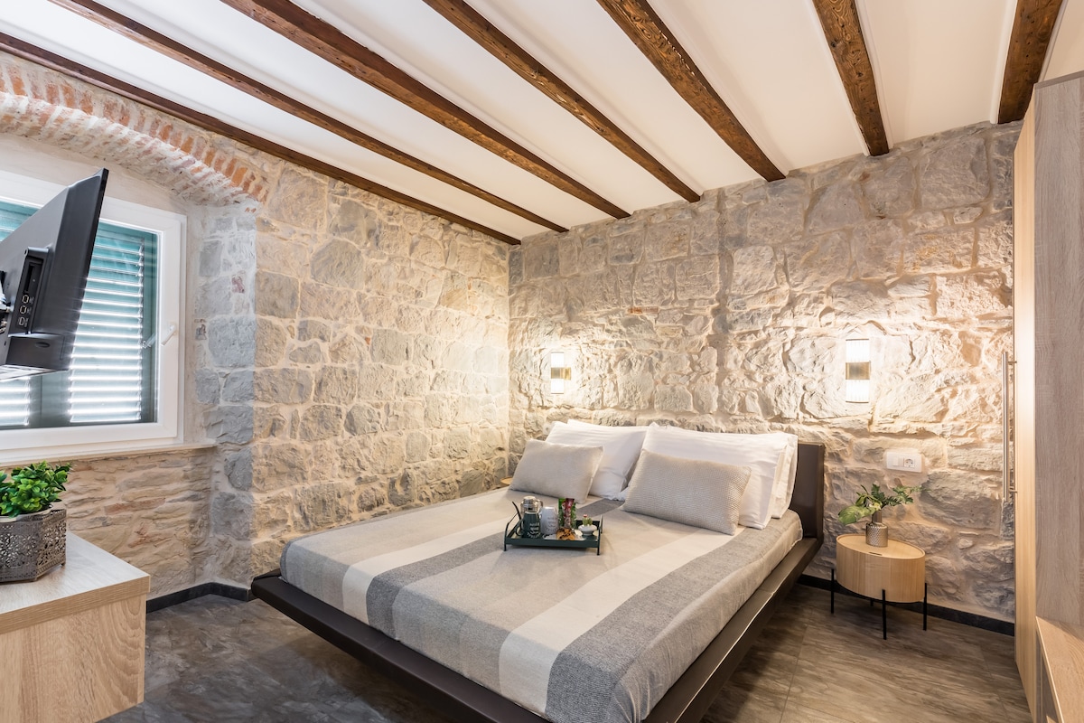A cozy bedroom is featured with a spacious bed dressed in neutral linens. Exposed stone walls provide a rustic touch, while wooden beams enhance the ceiling. A small side table holds a tray, and a television is mounted on the wall, adding to the room's functionality.