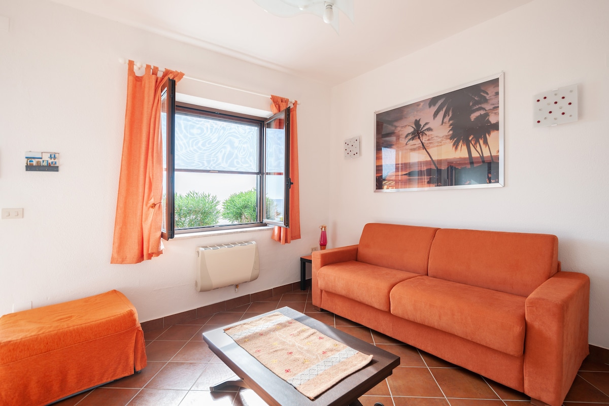 A cozy living room features an orange sofa and a matching pouf. A wooden table sits on the tiled floor, while bright curtains frame a window showcasing outdoor greenery. A colorful beach-themed artwork hangs on the wall, contributing to the inviting atmosphere.