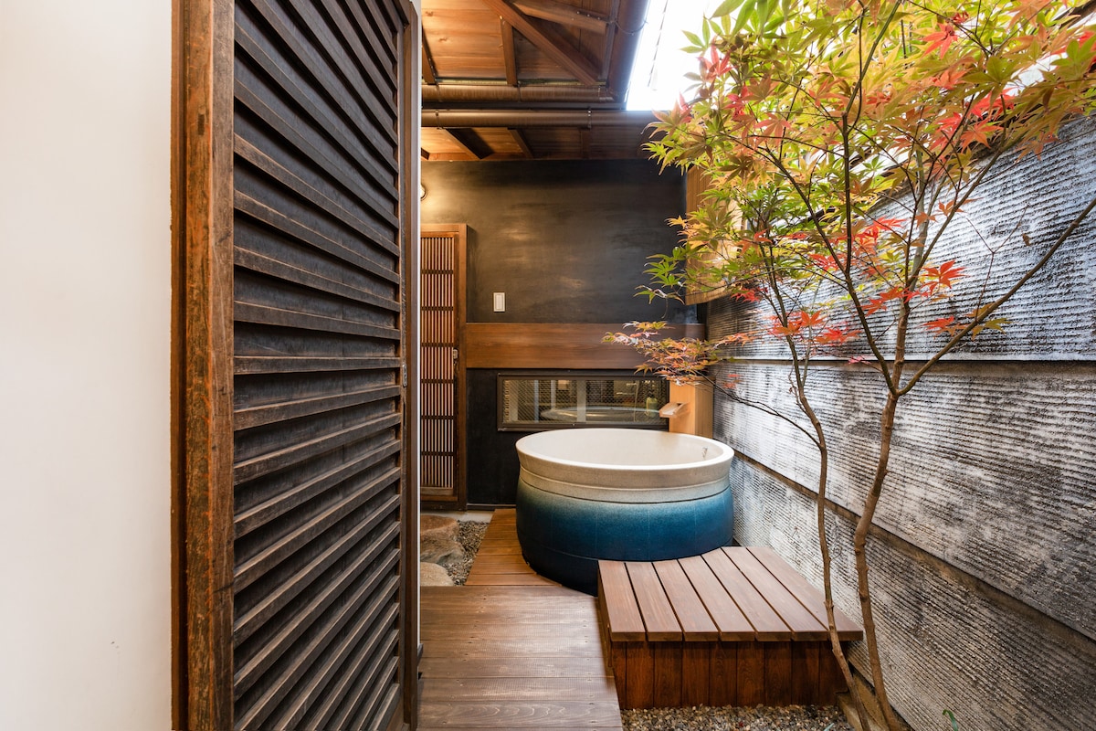 An outdoor space features a traditional Japanese ceramic bath surrounded by wooden decking and pebbled flooring. A vibrant tree adds greenery and a serene atmosphere, while textured wooden walls enhance the privacy and charm of the setting.