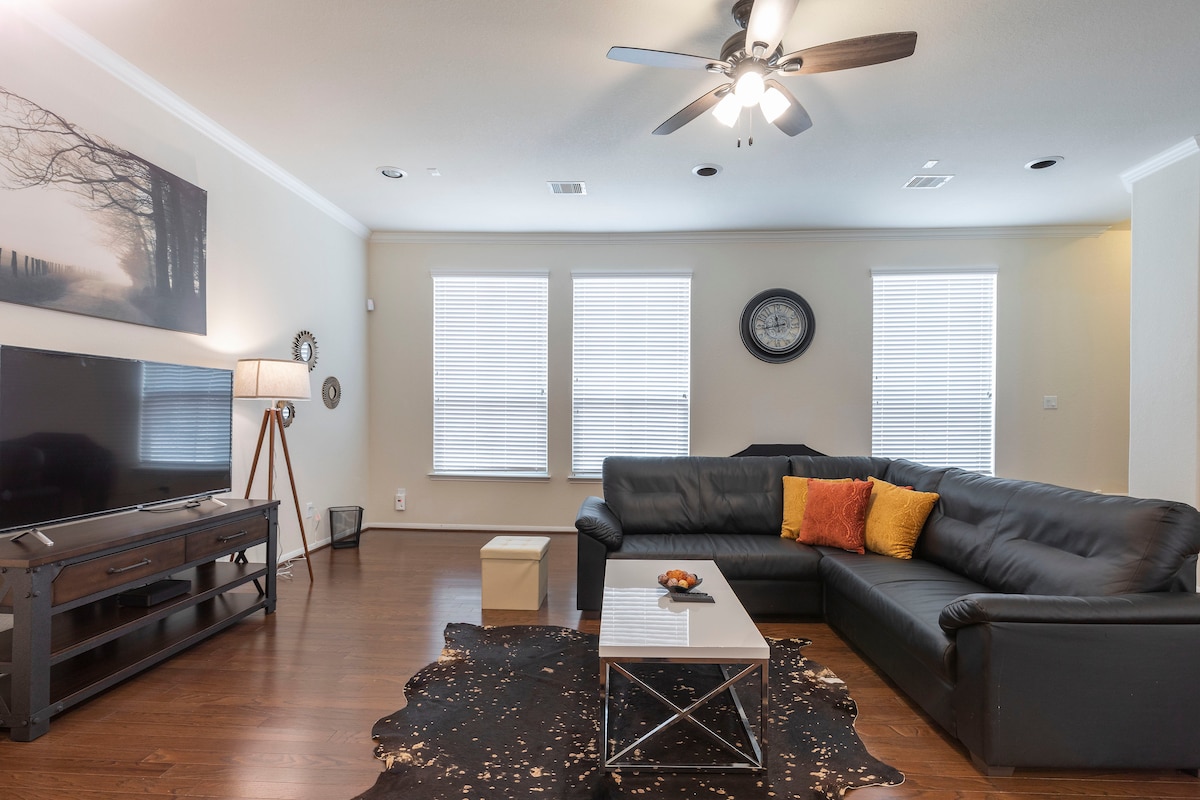 A spacious living area is equipped with a large sectional sofa and a sleek coffee table placed on a patterned rug. A television is mounted on an entertainment unit, while natural light enters through multiple large windows, enhancing the atmosphere.