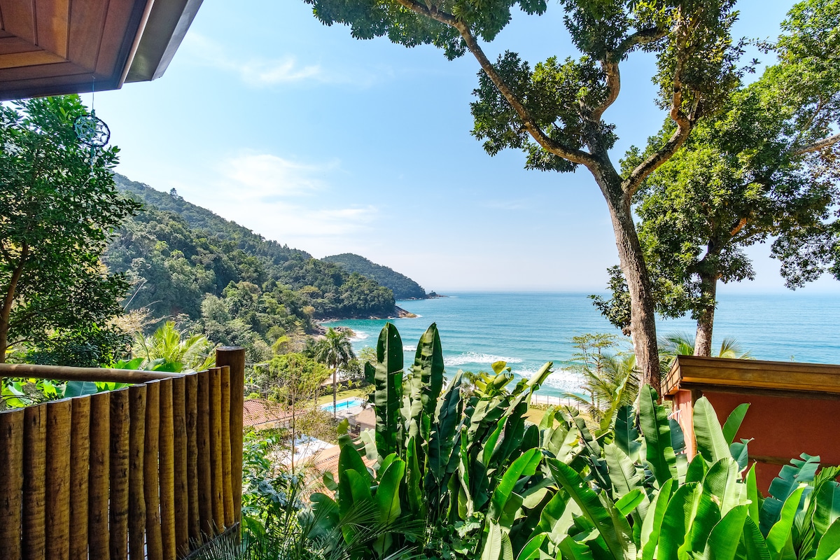 A scenic view from a wooden deck showcases the coastline with blue waters and lush greenery. Palm trees and tropical foliage frame the image, creating a serene natural environment. The distant hills provide a backdrop to the tranquil beachfront landscape.