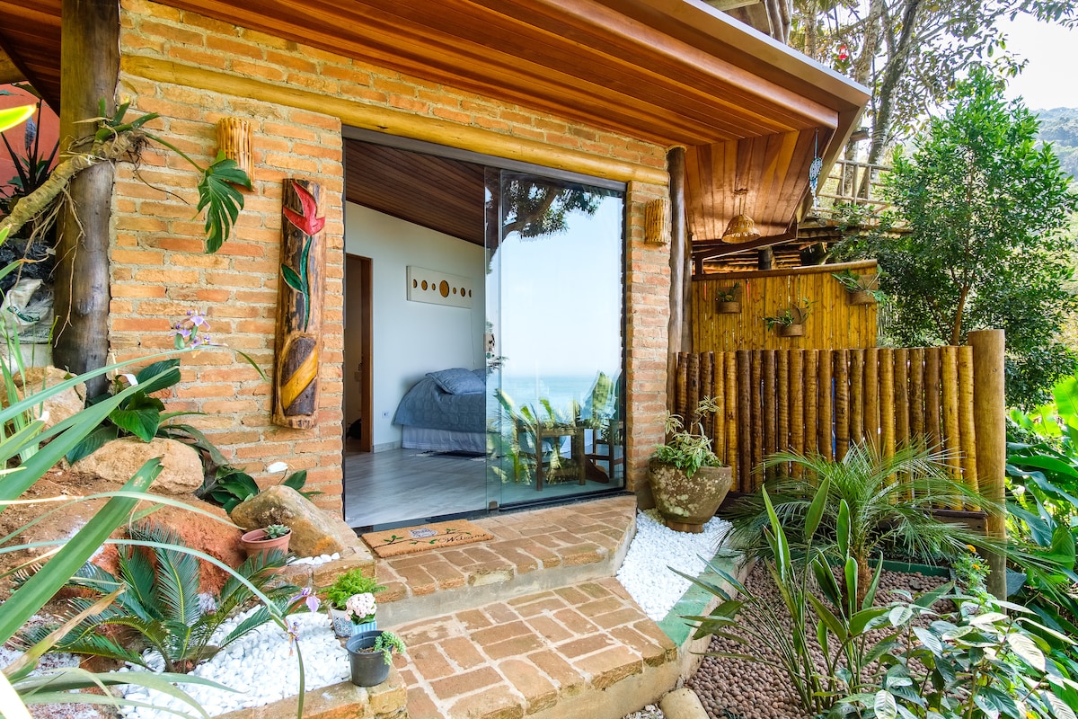 The exterior of the chalé features a rustic brick façade with a smooth glass door, opening to a nature-filled environment. Delicate plants and stones frame the entrance, while the nearby wooden fence complements the natural setting, enhancing the serene atmosphere of the property.