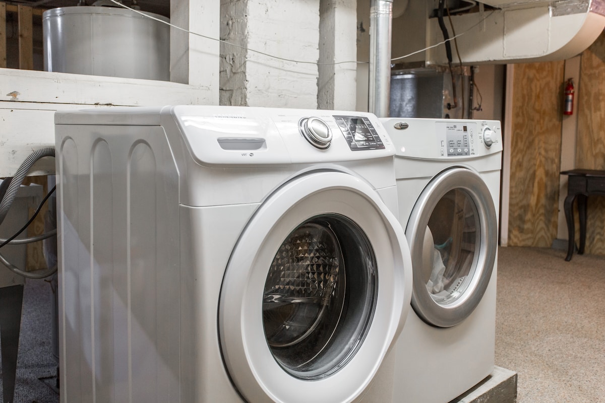 A pair of modern washing machines is positioned side by side in a well-lit basement area. The machines feature a sleek white design, with the dryer on the left and the washing machine on the right, both designed for convenient laundry use.