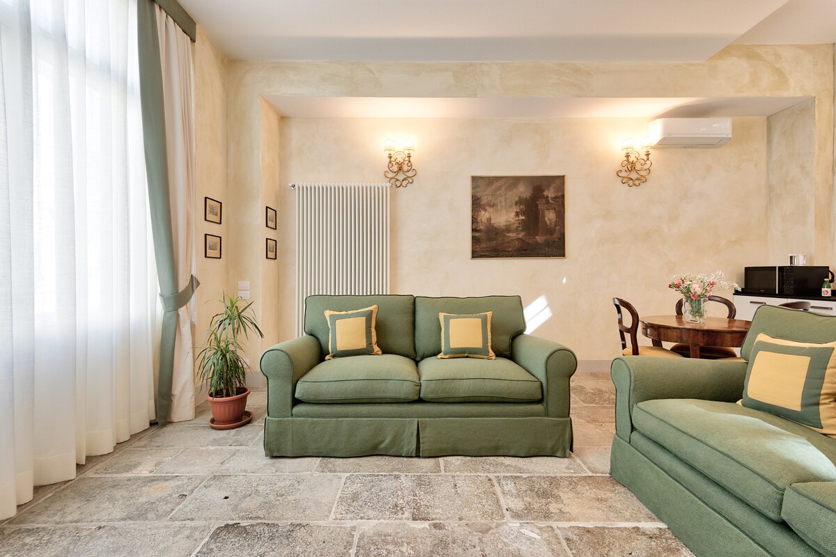 A living area is furnished with green sofas featuring yellow cushions. Natural light floods the room through large windows with sheer curtains. A dining table can be seen in the background, accompanied by a chair, while framed artwork adorns the walls.