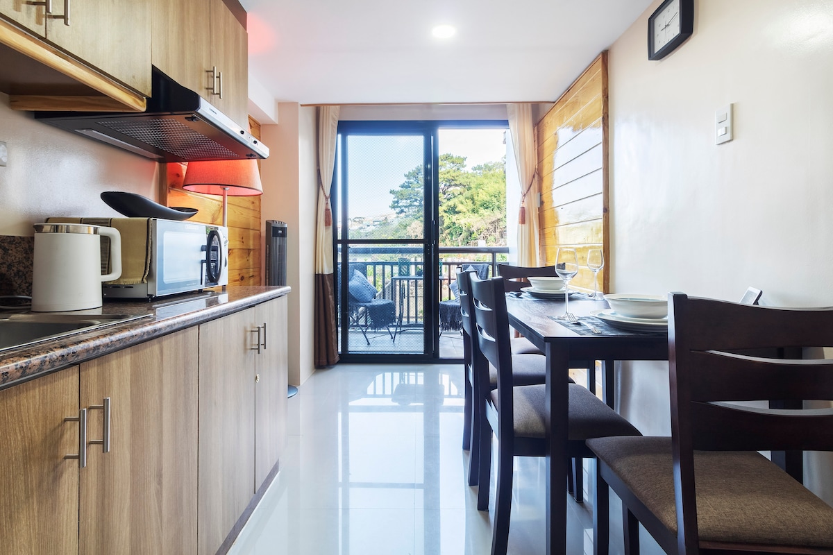 The modern kitchen area is equipped with wooden cabinetry and sleek countertops. A dining table is set against large glass doors that lead to a balcony. Natural light brightens the space, highlighting the cozy atmosphere with seating arrangements visible on the balcony.
