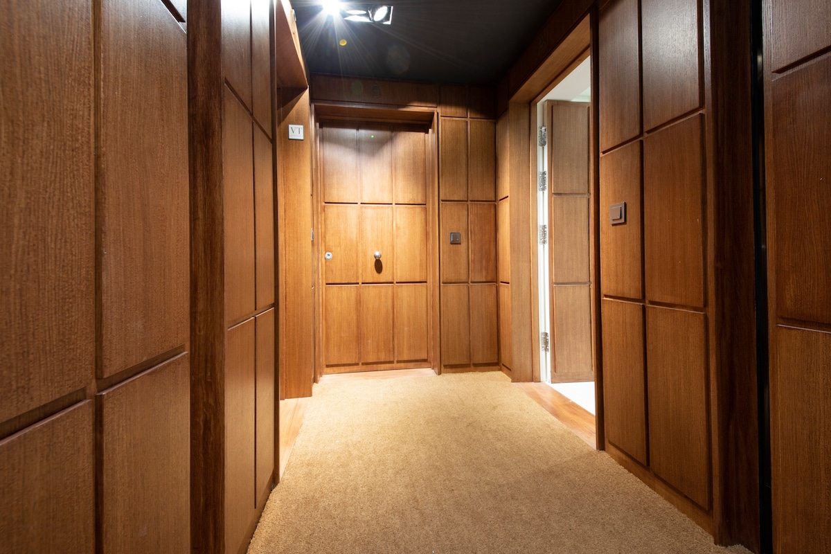 The hallway is lined with rich wooden paneling and features soft, neutral carpeting. A door is positioned at the end of the corridor, and there is a polished light fixture on the ceiling, providing illumination throughout the space.