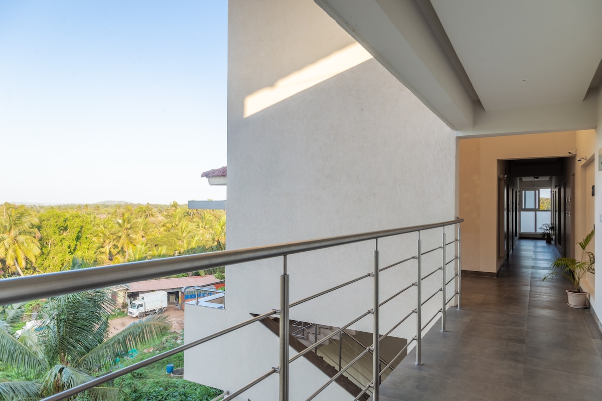 A breezy corridor is lined with sleek railings, showcasing a view of lush greenery and palm trees outside. The space features smooth, tiled flooring, and the warm sunlight creates a soothing ambiance. Doors lead to guest rooms, contributing to the inviting atmosphere.