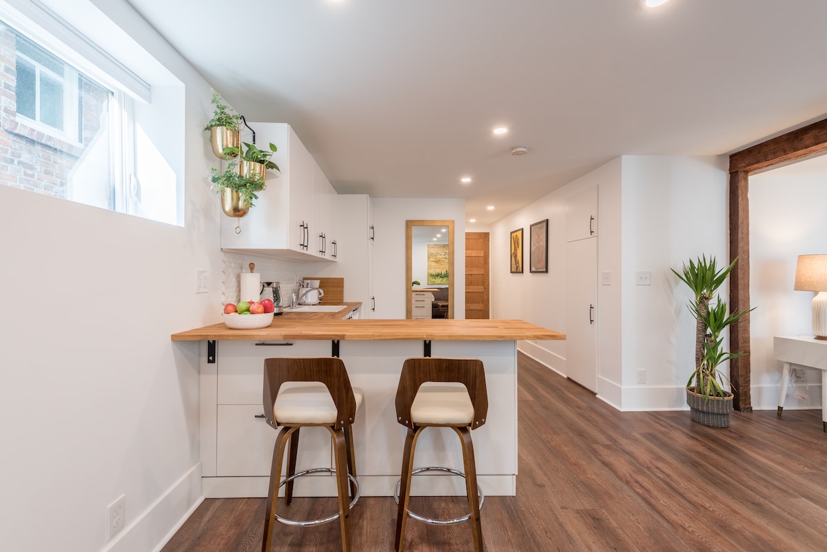 The kitchen features a minimalist design with a wooden breakfast bar and two high-backed stools. White cabinetry is complemented by a light-hued backsplash. A window lets in natural light, and a small plant adds a touch of greenery to the space.