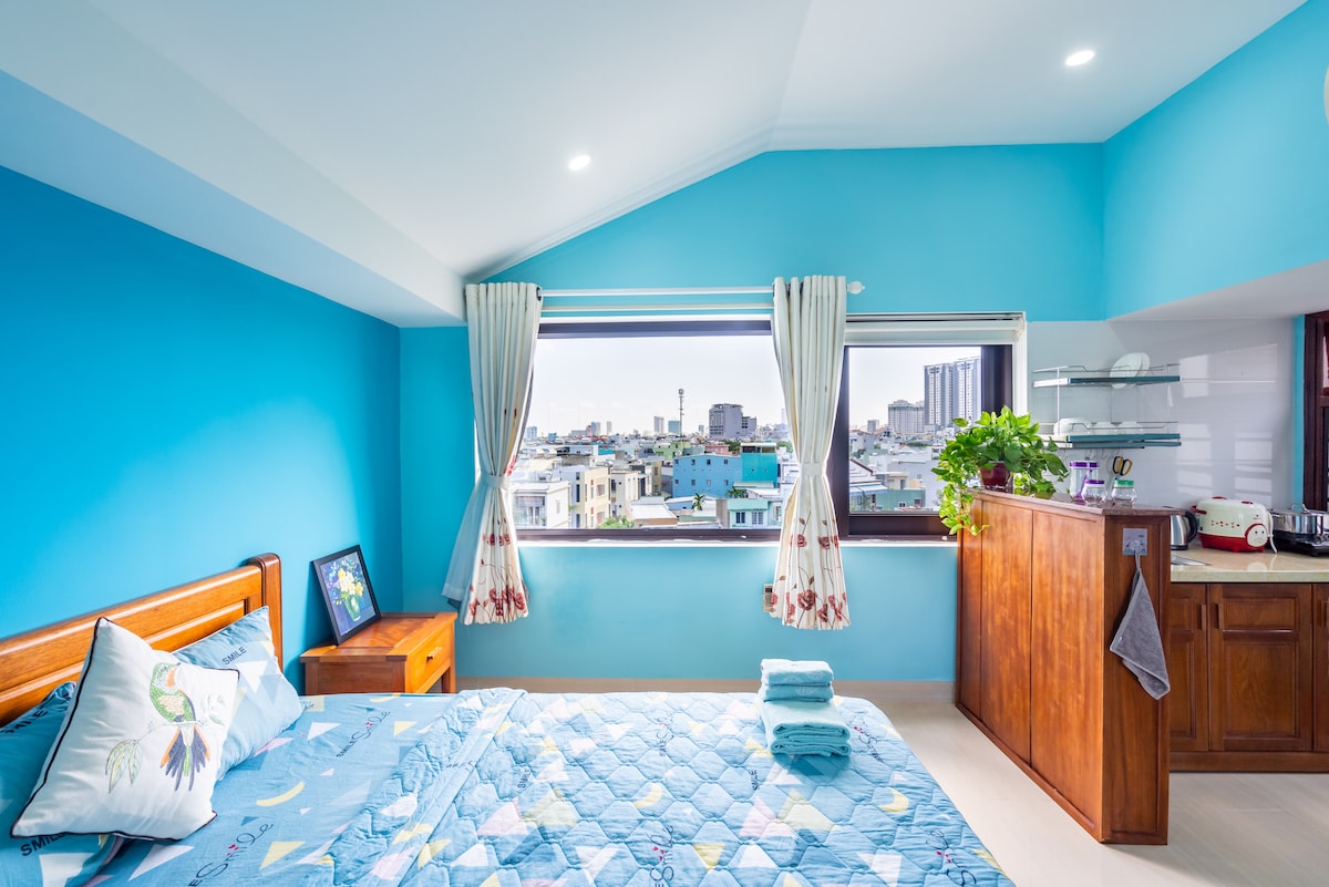 A bright bedroom features walls painted in soft blue with a quilted bedspread atop a spacious bed. Natural light floods the room through a large window that offers a cityscape view. A wooden nightstand and a kitchenette are visible, enhancing the room's functionality.