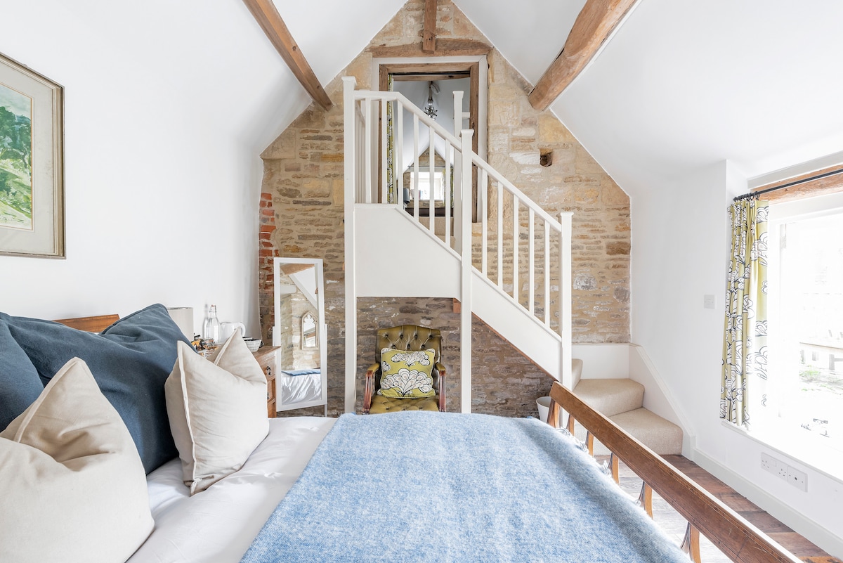 The vaulted bedroom features exposed stone walls and wooden beams. A spacious bed adorned with plush pillows is positioned in the foreground. A cozy seating area with a patterned chair is present, along with a staircase leading to the mezzanine level.