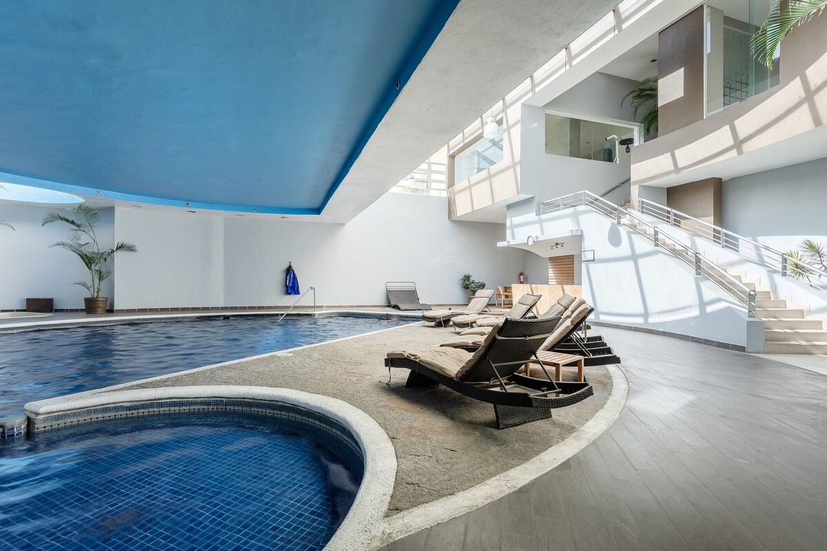 An indoor pool area features a spacious swimming pool with a jacuzzi nearby. Sun loungers are positioned around the pool, and large windows allow natural light to enhance the open space. Potted plants add a touch of greenery to the serene environment.