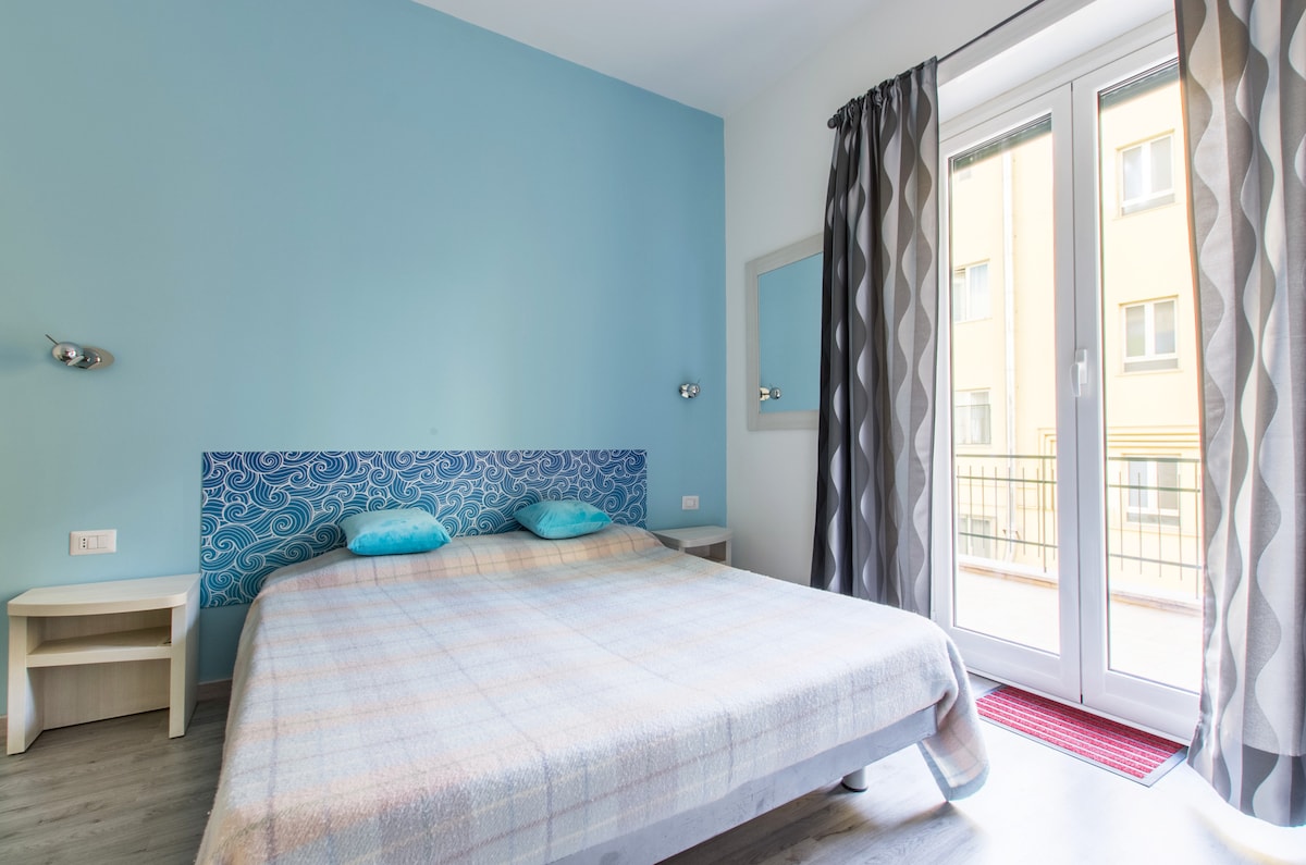 A spacious bedroom is equipped with a large bed dressed in neutral tones. Light blue walls complement a patterned headboard. Natural light filters through large double doors that open to a small balcony, accentuated by floor-to-ceiling curtains in a modern design.