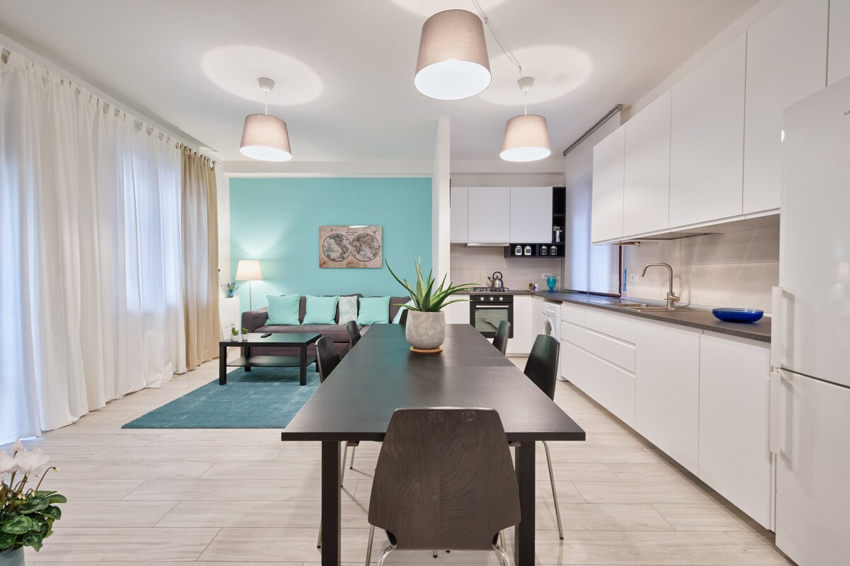 An open-plan living area is featured, with a dining table for six and a kitchen in the background. Soft light is provided by pendant lamps above. A comfortable seating arrangement is positioned next to a bright blue wall, complemented by a large area rug beneath.