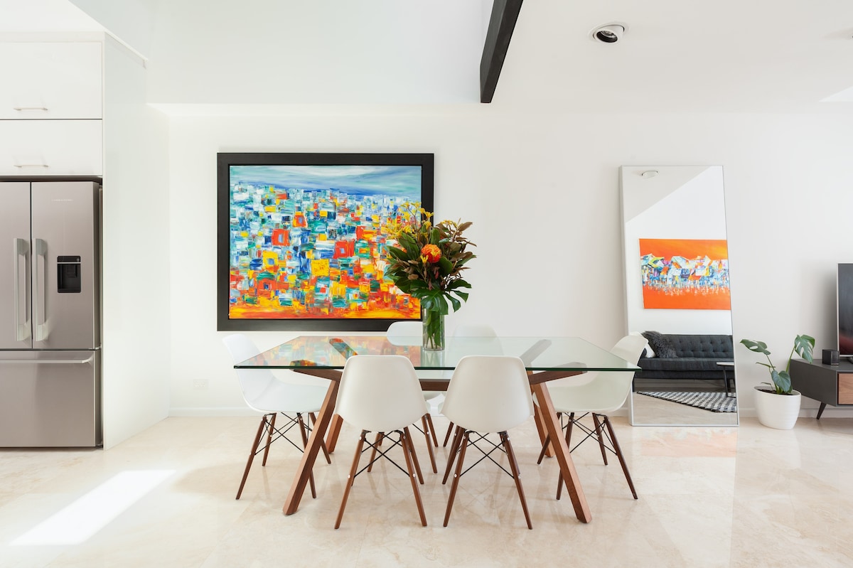 A modern dining area features a glass table surrounded by six stylish chairs. A large, colorful artwork occupies the wall, adding a splash of vibrancy to the room. Natural light floods in, illuminating the polished tile floor, while a black sofa and mirror are positioned against the opposite wall.