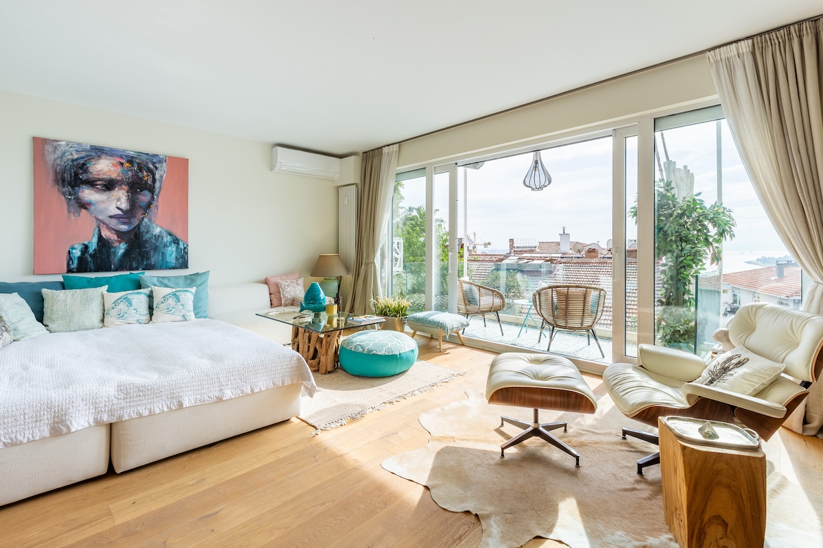 A spacious living area is featured, showcasing a large bed adorned with layered textiles. A glass coffee table sits at the center, surrounded by comfortable seating. Natural light pours in through expansive windows, which lead to a balcony offering urban views. Greenery adds a refreshing touch.