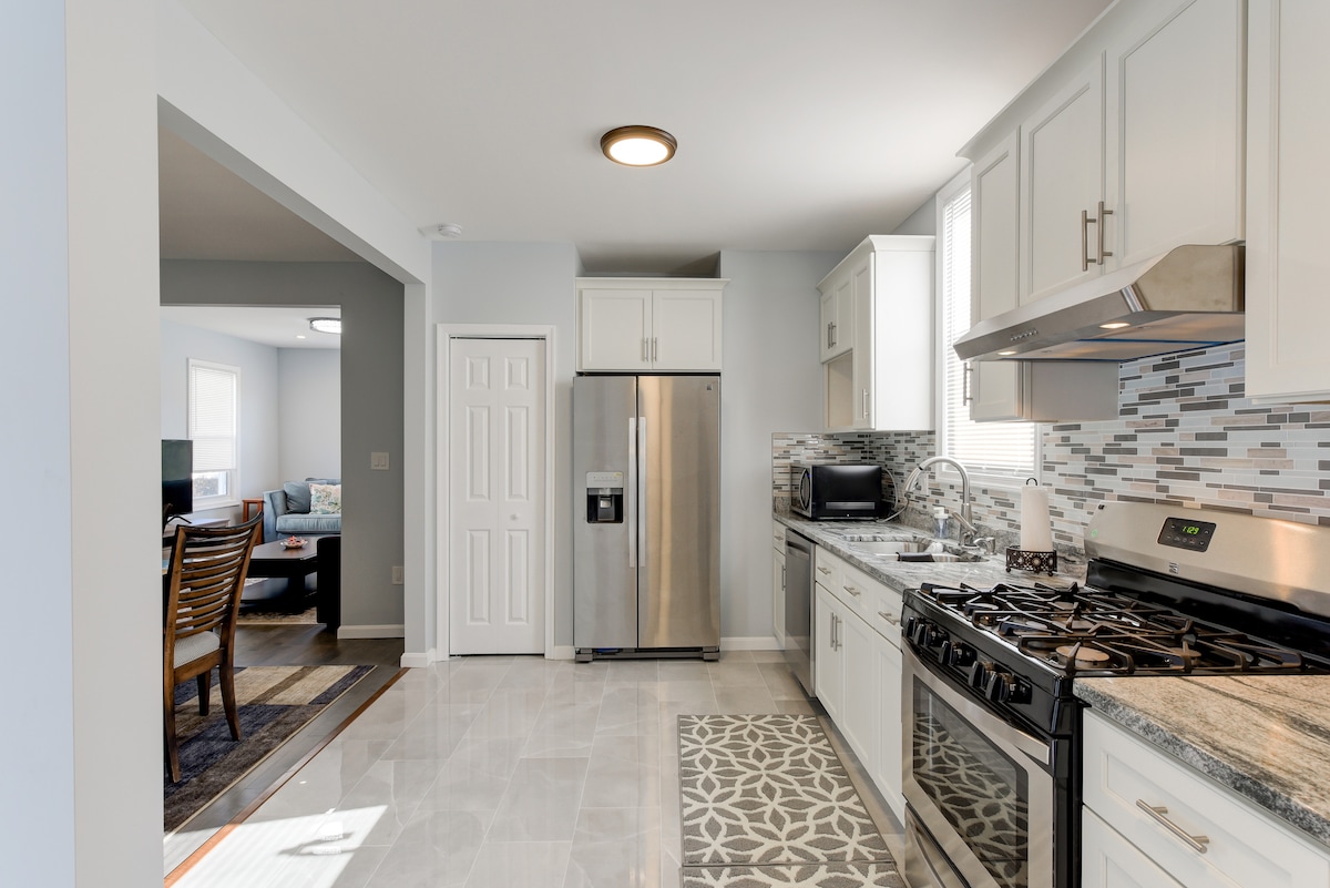 A well-equipped kitchen features modern stainless steel appliances, including a refrigerator and stove. Light-colored cabinetry and a patterned floor enhance the space. A doorway leads to a living area, providing an open and welcoming layout filled with natural light.