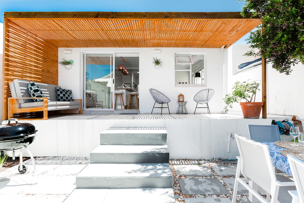 A modern outdoor area features a wooden patio covered by a slatted roof, providing shade. Comfortable lounge chairs are positioned on the patio, alongside a dining table set for guests. A barbecue grill is nearby, and steps lead down to a stone pathway surrounded by greenery.
