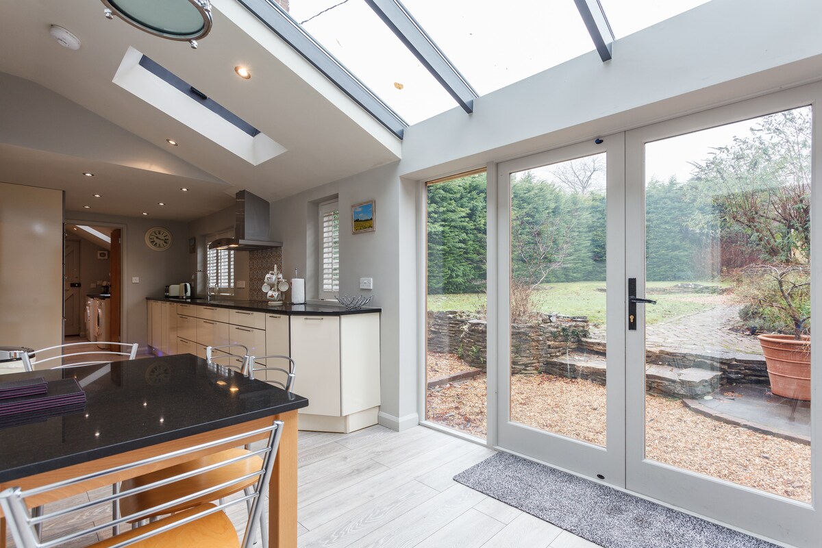 A modern kitchen featuring cream cabinetry and granite work surfaces is illuminated by natural light from the glazed roof above. A breakfast table with seating is positioned near the sliding glass doors, which open to a serene garden area surrounded by greenery.