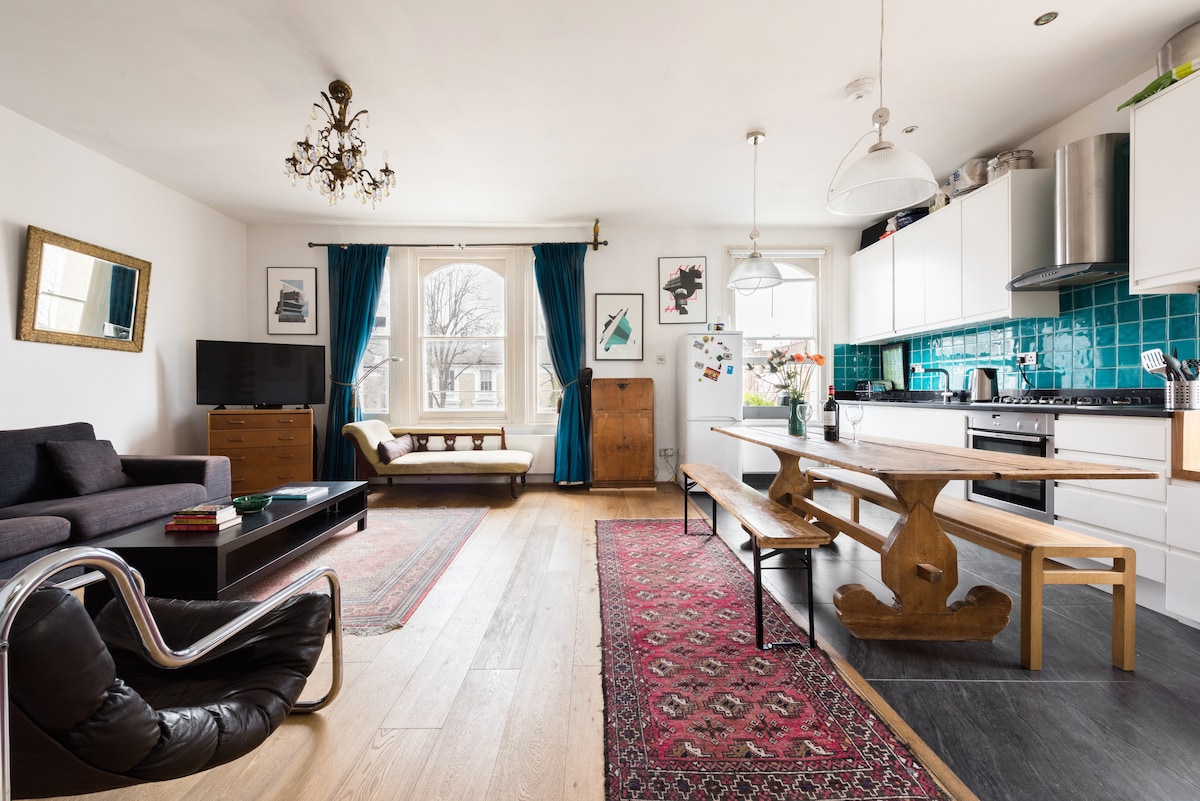 An open-plan living area features large windows with floor-length curtains, a wooden dining table with benches, and a comfortable sofa facing a smart TV. Decorative artwork and a patterned rug add character to the room, complemented by modern kitchen appliances and stylish light fixtures.