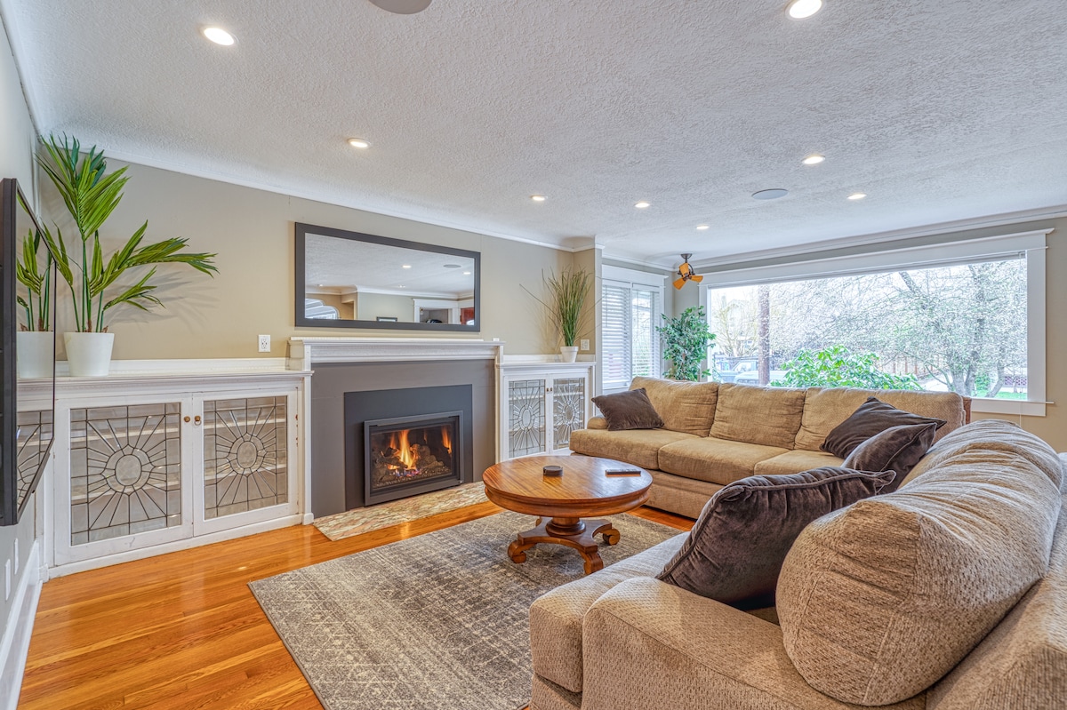 A spacious living area is highlighted by a large sectional sofa and a central coffee table. A cozy gas fireplace is set within a modern mantle, framed by stylish decor. Natural light floods the room through expansive windows, accentuating the hardwood floors and soft area rug.