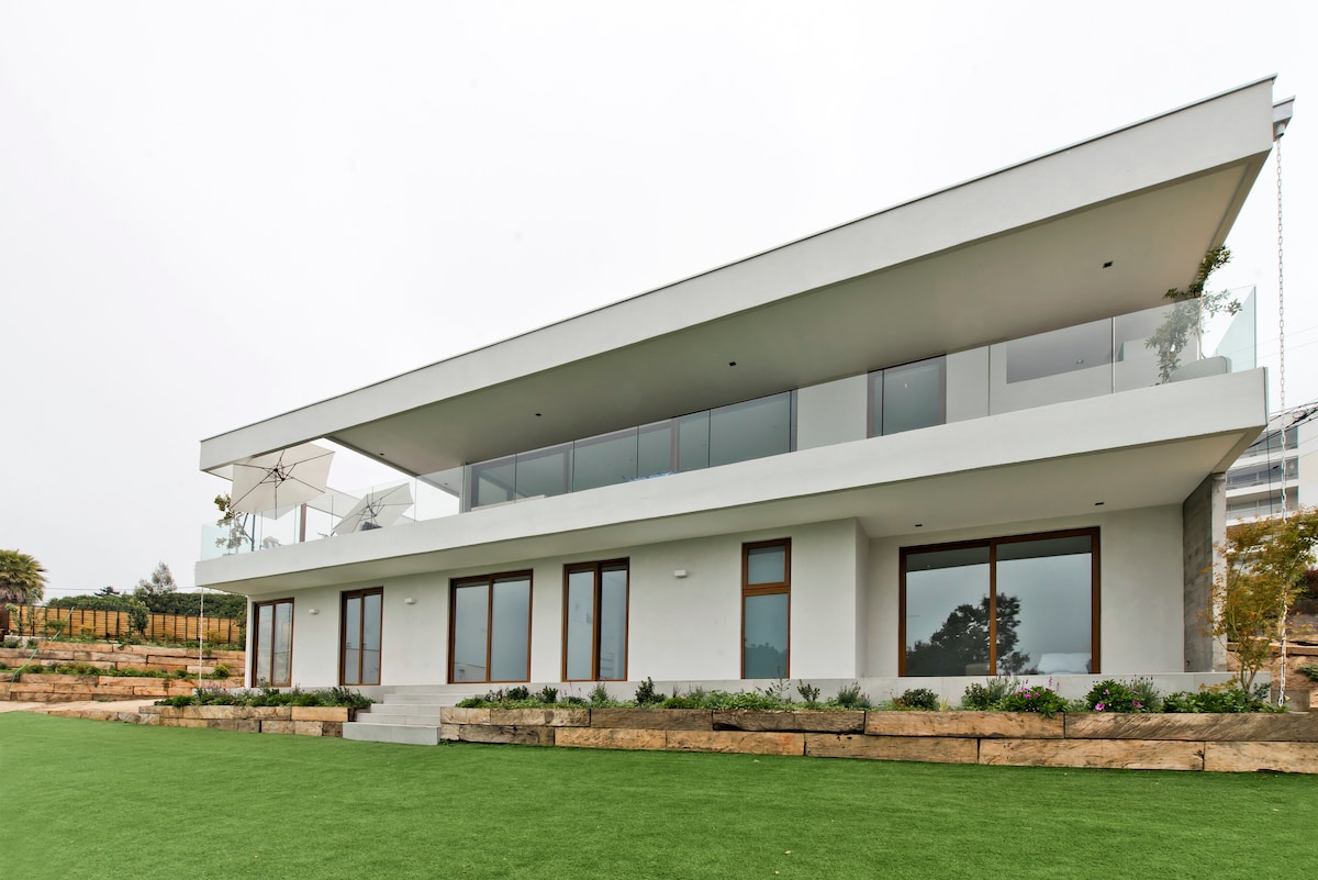 A modern two-story house is showcased with a clean white exterior. Large glass windows are visible on both levels, allowing natural light to fill the interior. A spacious lawn surrounds the property, complemented by neatly arranged flower beds along the base.