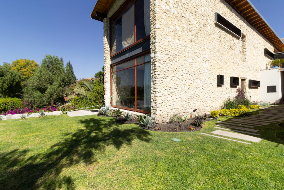 The exterior of a modern stone house is showcased, featuring large windows that invite natural light. A well-maintained garden surrounds the property, with vibrant flowers and greenery complementing the structure. A pathway leads to the entrance, enhancing the inviting atmosphere of the setting.