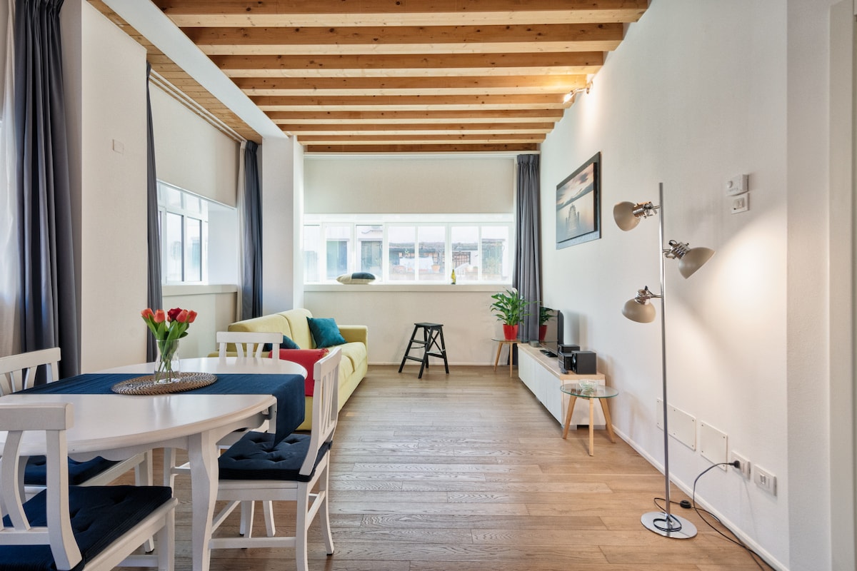 A comfortable living space features wooden beams overhead, with a yellow sofa and a small black stool. A dining table with chairs sits adjacent, adorned with a vase of tulips. Natural light filters through large windows, brightening the interior.