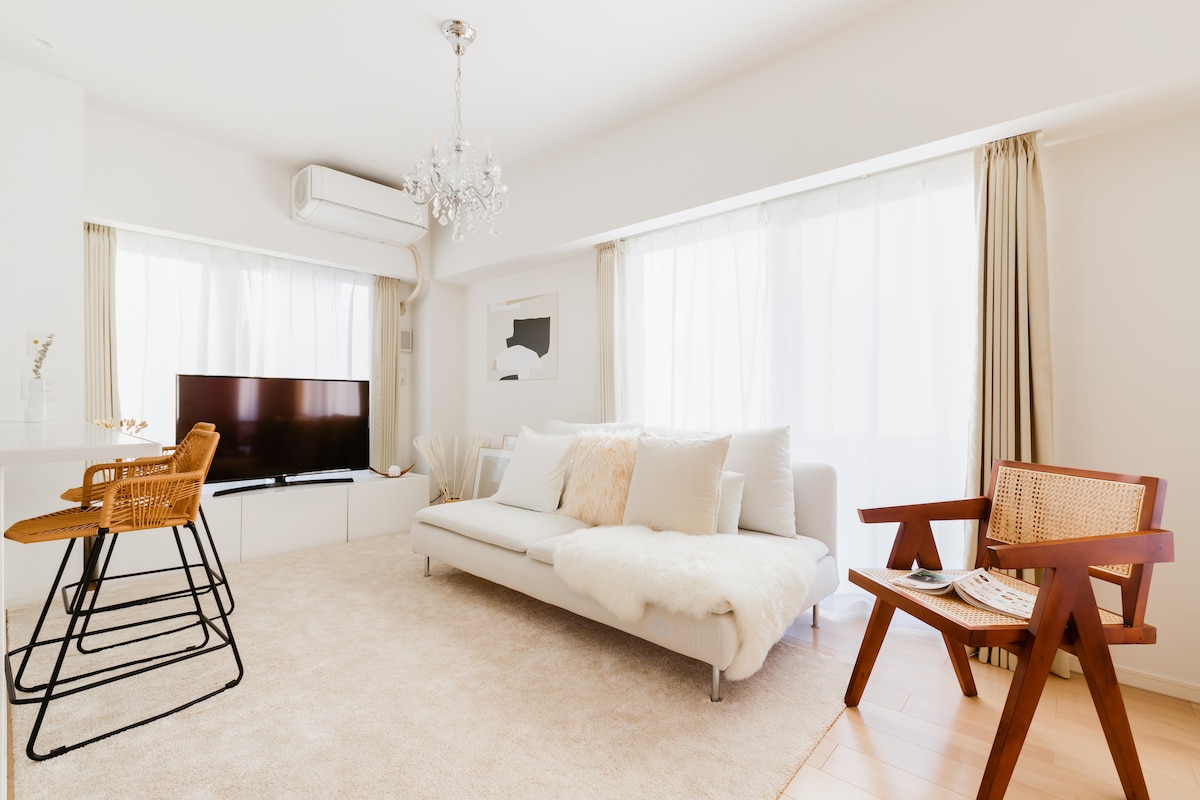 A light-filled living area features large windows dressed with sheer curtains, creating an airy ambiance. A comfortable sofa is complemented by a decorative throw, while a TV is positioned on a white cabinet. A stylish armchair and sleek bar stools add to the inviting decor.