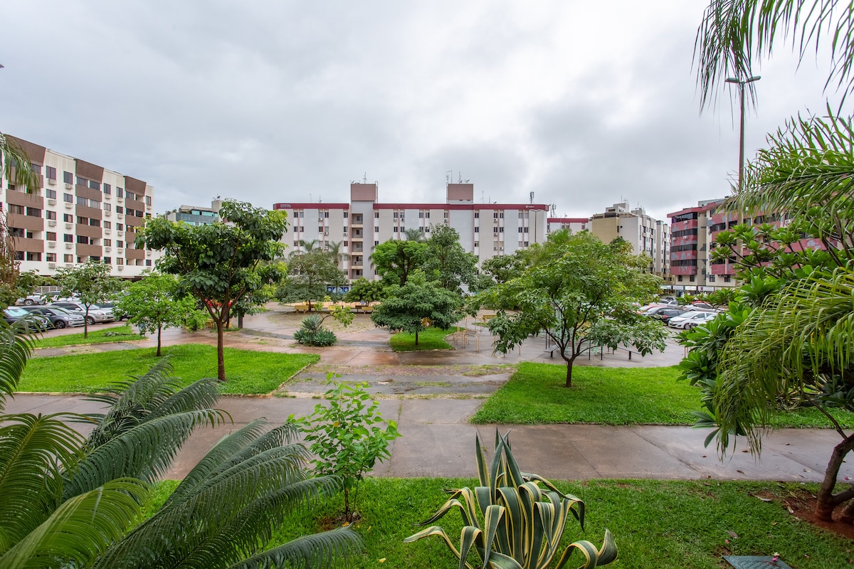 A view of the residential complex and its surroundings features well-maintained green spaces with trees and grass. Parked cars are visible along the pathways, and the buildings' facades create a structured backdrop. The scene is set under an overcast sky.