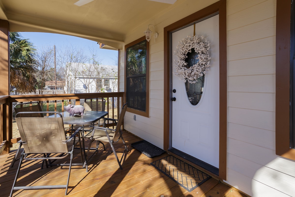 A welcoming porch area features a wooden deck with a table and four chairs. A decorative wreath enhances the entrance door, while natural light filters through the surrounding windows. Lush greenery is visible in the outside garden area, providing a serene backdrop.