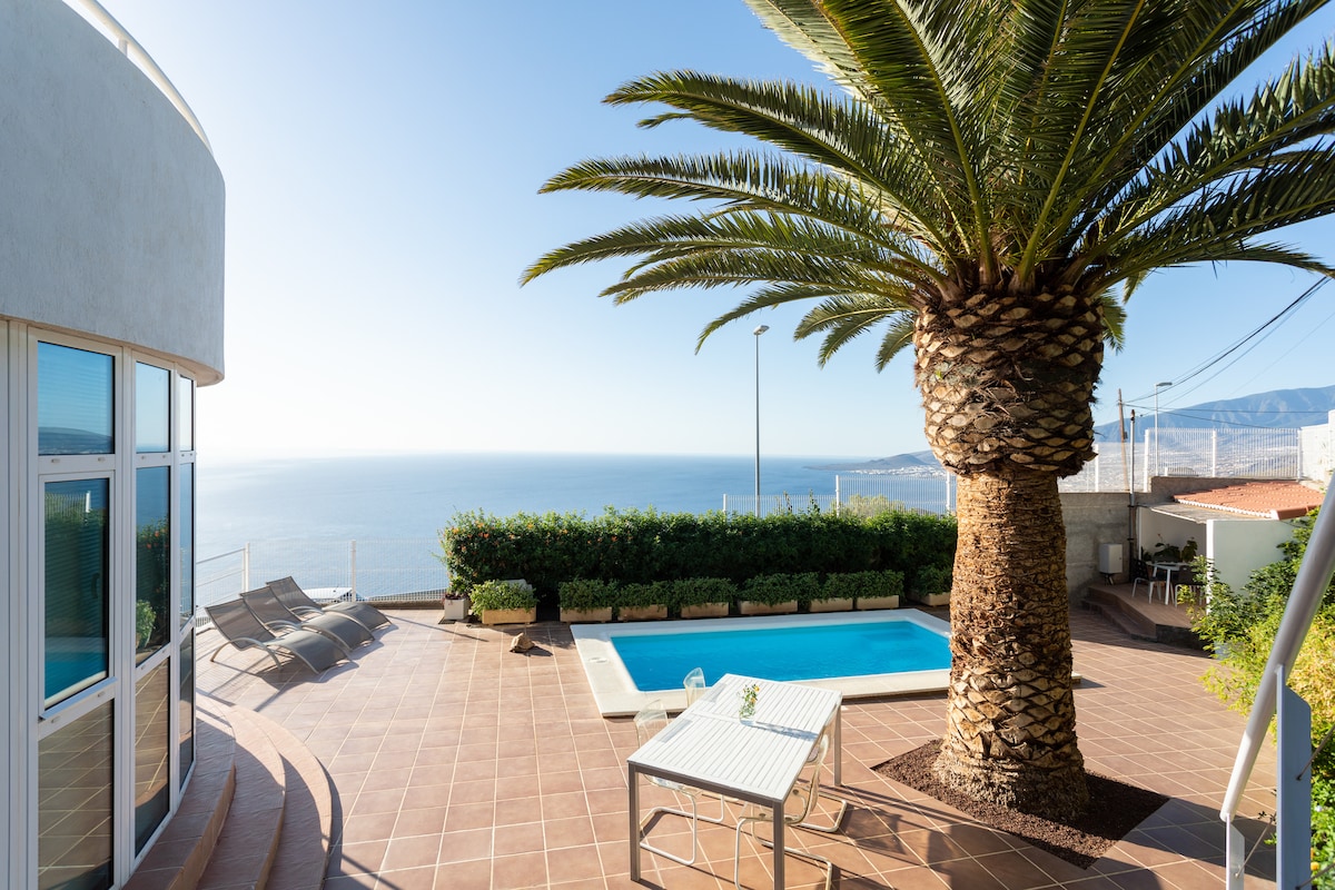 An outdoor space featuring a pool surrounded by a tiled terrace. A tall palm tree provides shade, while loungers are positioned nearby. The view overlooks the ocean, creating a serene atmosphere in a well-maintained area with greenery in the background.