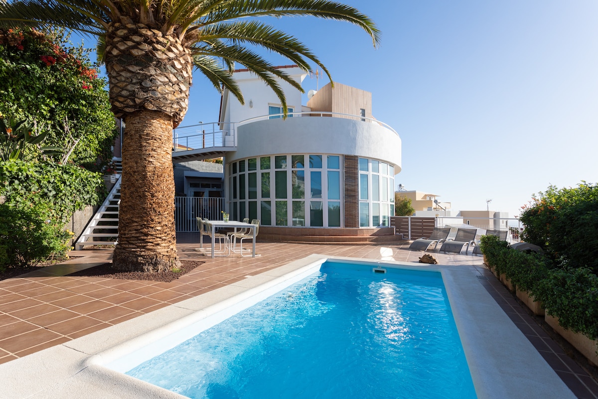 An outdoor space is presented with a sparkling swimming pool surrounded by tiled decking. Lush greenery, including palm trees, enhances the atmosphere. The house, featuring large windows, is complemented by a small dining table and chairs situated nearby.