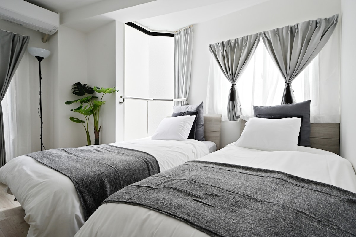 The room features two spacious beds with neutral-colored bedding and decorative gray throws. Natural light fills the space through large windows dressed in elegant curtains. A small plant adds a touch of greenery to the corner, enhancing the calming atmosphere.